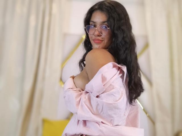 CloverLover with long black wavy hair, pink glasses, and pink shirt off-shoulder, covering her tits, smiling at the camera. Soft pink and yellow background.