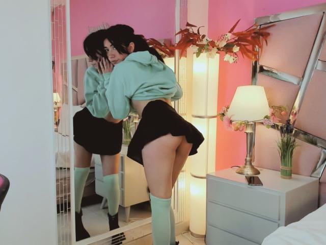 AnyuWu with her ass up, wearing a green hoodie, black skirt, and green knee socks, reflected in a mirror, showing her bare ass. Bedroom with pink walls.