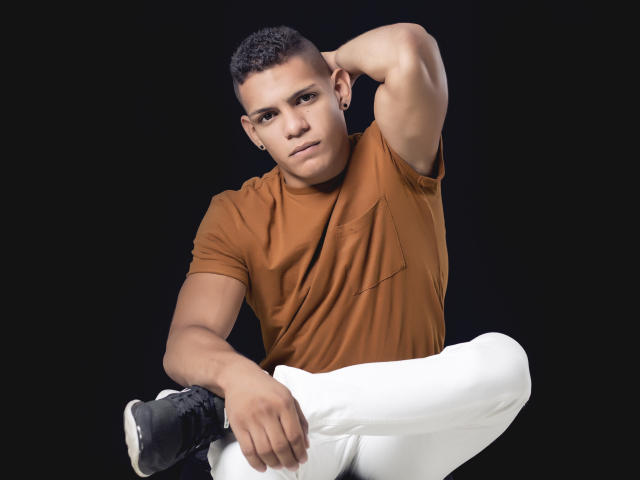 LoganVelasco with short curly black hair, brown t-shirt, white pants, black shoes, muscular arms, ear piercings, black background, arm raised, hand behind head, looking at camera.