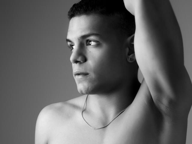 LoganVelasco is shirtless with a chain necklace, raising his right arm. He has short dark hair, a small mustache, and a focused expression. Black-and-white, head-and-shoulders shot.
