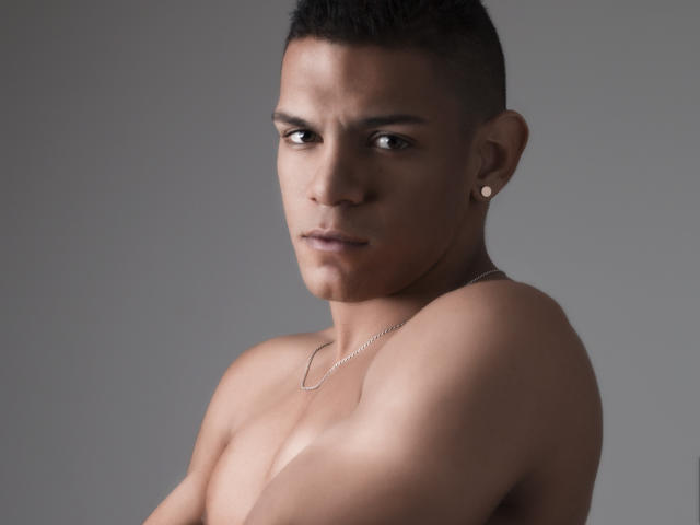 LoganVelasco, shirtless, has a clean-shaven face, short black hair, a small earring in his left ear, and a silver necklace. His eyes are serious, looking slightly to the side.