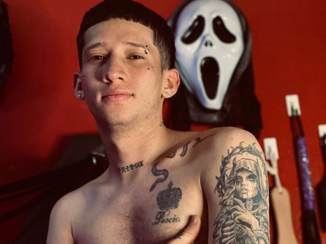 Shirtless AlexAndAlex with short black hair, ear piercings, and tattoos on chest and arm, holding his tits, standing in front of a white mask. Red wall background.