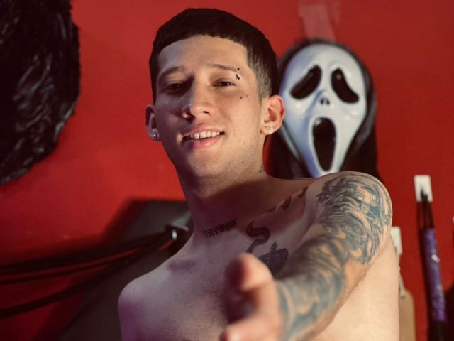 Shirtless AlexAndAlex with tattoos, short black hair, nose ring, and earrings, pointing forward, smiling, red wall, white masked figure in background.