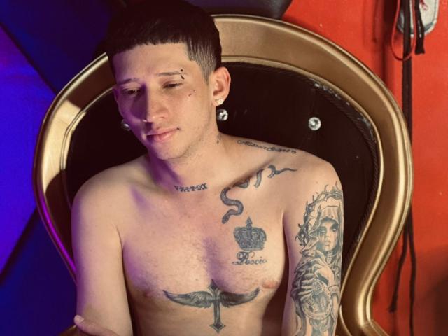 Shirtless AlexAndAlexa with short dark hair, ear piercings, tattoos, and a crown tattoo on chest, sitting in a gold chair, looking slightly to the side with a slight smile.