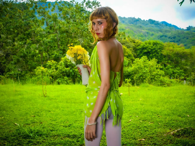 AuraFire with short brown hair, green leaf dress, white polka dots, holding yellow flowers, standing in grassy field, looking back, wearing white pants, mountain forest in background.