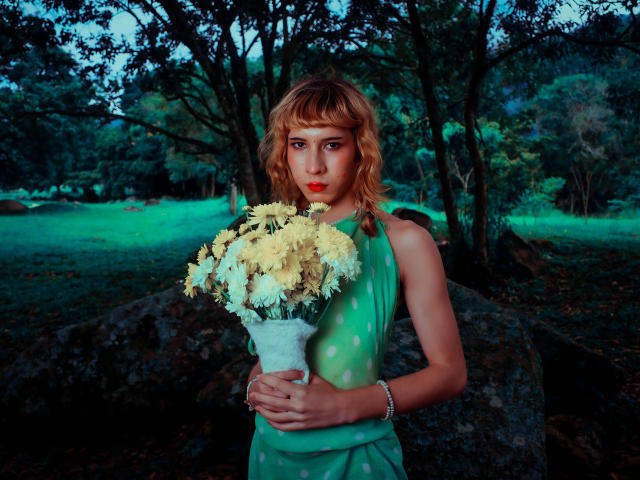 AuraFire with shoulder-length blonde hair, wearing a green polka-dot dress, holding a bouquet of white flowers, standing in front of a tree, dusk light, serious expression, red lipstick.