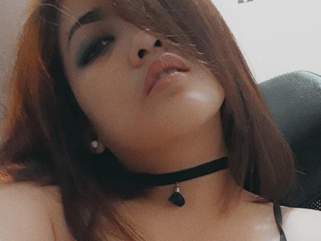 CherryRedForYou has brown hair, dark smoky eye makeup, glossy pink lips, black choker with heart pendant, and is looking up with a slight pout. Boobs not visible.