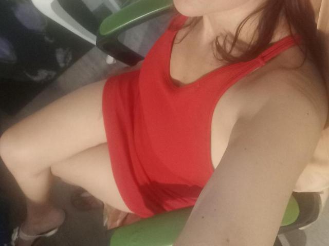SherahaJade trägt ein rotes Tanktop und Shorts, sitzt mit verschränkten Beinen, wobei ihre nackten Arme sichtbar sind und ihre Brüste und ihr Hintern zu sehen sind. Ihre roten Haare sind sichtbar..