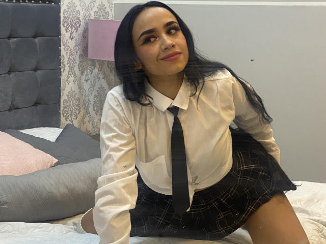 MacarenaColleman with long black hair, wearing a white shirt, black tie, and plaid skirt, sitting on a bed, smiling, showing her ass slightly, looking cute.