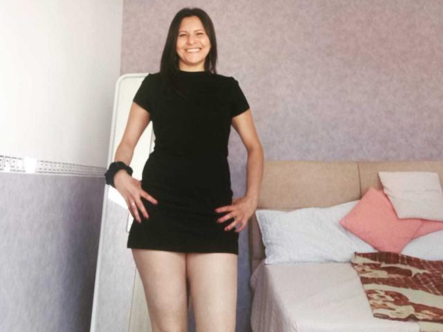 VeronikaGreco è in piedi, sorridente, indossa un vestito nero aderente, ha le mani sui fianchi, mostrando il sedere. Ha i capelli neri, un elastico nero al polso e un cuscino rosa..