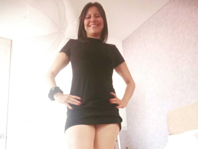 VeronikaGreco, con seno di media grandezza, vestito nero, mani sui fianchi, sorridente, braccialetto nero, capelli castani corti, in piedi in una stanza luminosa, glutei visibili, unghie rosse, pelle chiara, corporatura media..