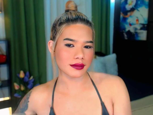 XFullOfLoadTransX, met blond haar in een knot, draagt een zwarte haltertop, heeft roze lippenstift, donkere eyeliner en een tatoeage op haar rechter schouder, en kijkt naar de camera..