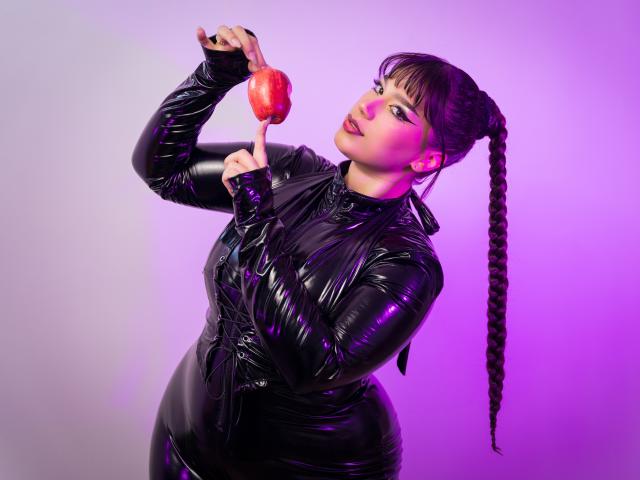 Milly Johnson, com longos cabelos negros trançados, vestindo um vestido preto de látex brilhante, segura uma maçã vermelha perto do nariz, sob iluminação roxa e rosa..