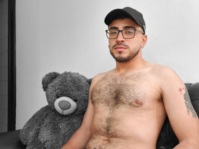 Shirtless DanteSexy with a hairy chest, black cap, glasses, and a tattoo, sitting next to a large gray teddy bear, looking at the camera. DanteSexy's dick is not visible.