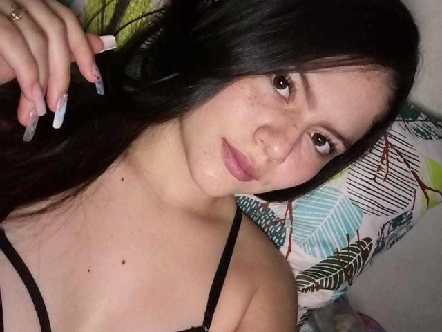 Sukilove has pale skin, dark long hair, and is lying down with a slight smile. She's wearing a black strap top, showing small tits. Her nails are long and white.