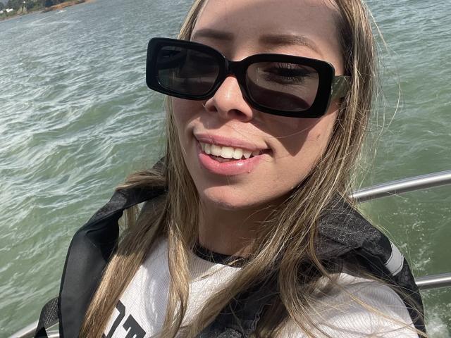 JennyLawry's face with black sunglasses, blonde hair, and a white shirt, smiling with pink lips. She's wearing a black jacket and standing on a boat with water.