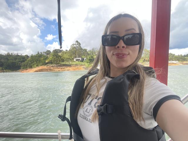 JennyLawry with medium tits, wearing black sunglasses, white shirt, and black life jacket, smiling at the camera from a boat, with green water and forested island in background.
