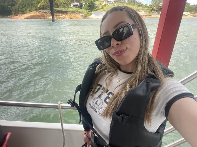 JennyLawry with sunglasses, smiling, wearing a black life vest over a white shirt, standing on a boat, green water in background, red pole on right.
