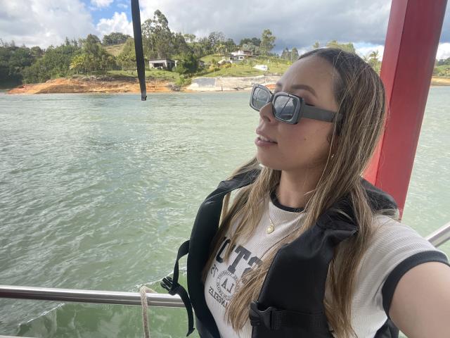 JennyLawry, con el cabello castaño mojado, gafas de sol negras, camisa blanca, chaleco salvavidas negro y un pequeño collar dorado, de pie en un bote, mirando hacia un lado, cerca de una columna roja, en agua verde..