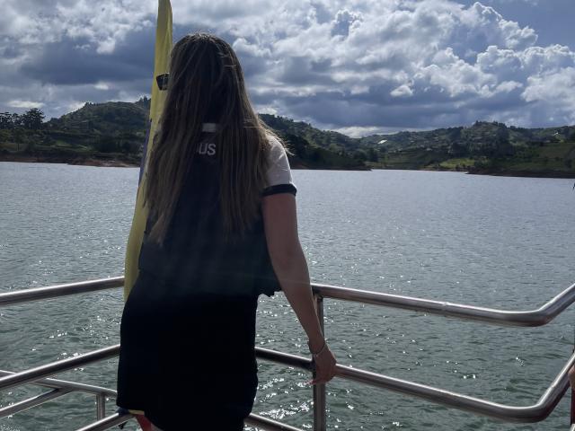 JennyLawry met lang bruin haar, een zwart-wit shirt, staat op een boot, met haar rug naar de camera gericht, en houdt een geel surfboard vast, boven kalm water, onder een bewolkte lucht..