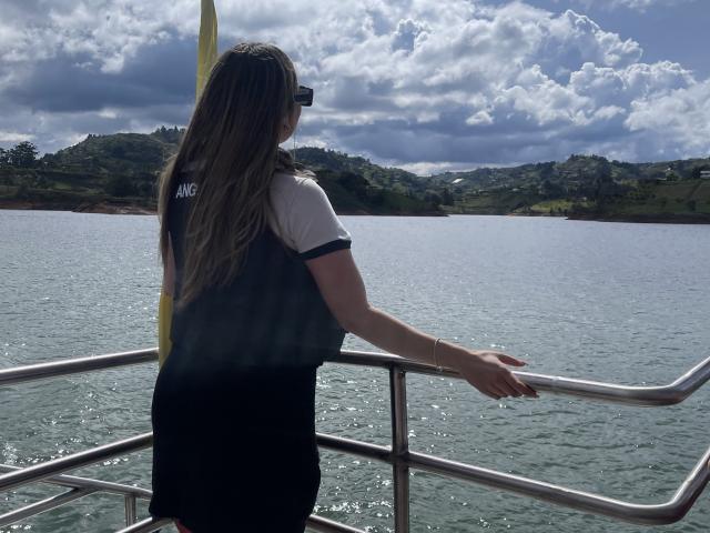 JennyLawry, avec ses longs cheveux bruns, portant une chemise noire et blanche, se tient sur un bateau, les bras appuyés sur une rambarde en métal, face à un ciel bleu et nuageux au-dessus d'une voie navigable verdoyante..
