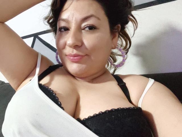 SakotoDika, con cabello oscuro, vestida con una camiseta blanca y un sujetador de encaje negro, mostrando sus grandes senos, aros morados y una ligera sonrisa, con el brazo levantado..
