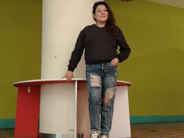 SakotoDika with long brown hair, wearing a black sweater and ripped blue jeans, leaning against a white and red cylindrical structure, smiling slightly.