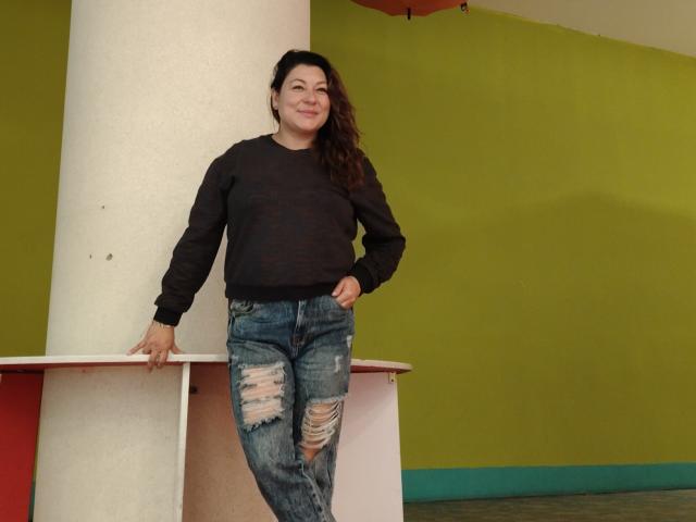 SakotoDika, com cabelos castanhos longos, suéter preto e jeans azuis rasgados, apoiada em um pilar branco, sorrindo, com as mãos nos bolsos, com uma aparência casual e relaxada..