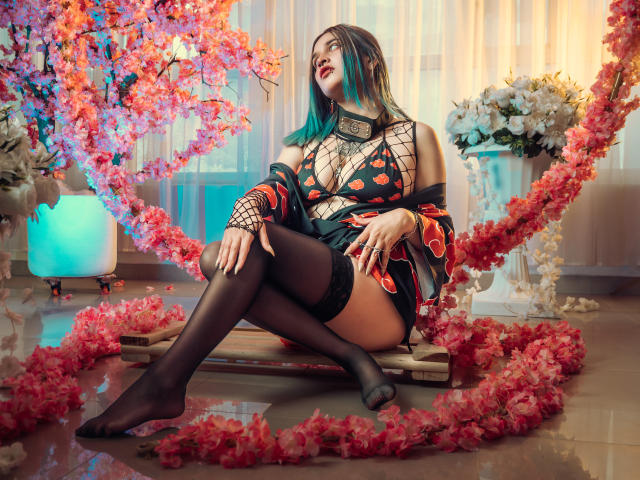 MaiaThunder with blue-green hair, black fishnet top, red-orange pattern, black thigh-highs, and black shorts, sitting on wooden planks, surrounded by pink flowers, and white roses.