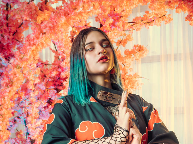 MaiaThunder with teal hair, black ninja outfit, fishnet sleeves, and metal collar, looking smug, hands up, cherry blossom background, red lipstick, nose ring, and blue eye makeup.