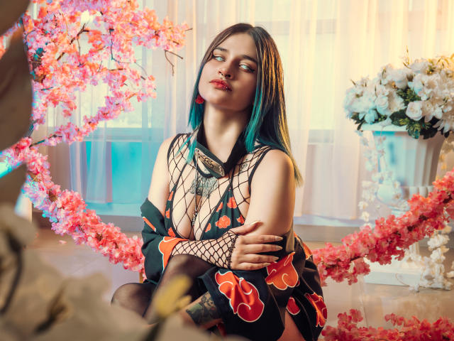 MaiaThunder with blue-green hair, wearing a fishnet top, revealing her tits, and a black dress with red butterfly patterns, sitting amid pink cherry blossoms, looking to the side.