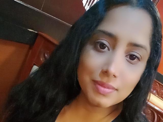 MariaCatalina's face with medium brown skin, long black wavy hair, and pink lips, showing a slight smile, with brown eyes and subtle makeup, in a casual selfie.