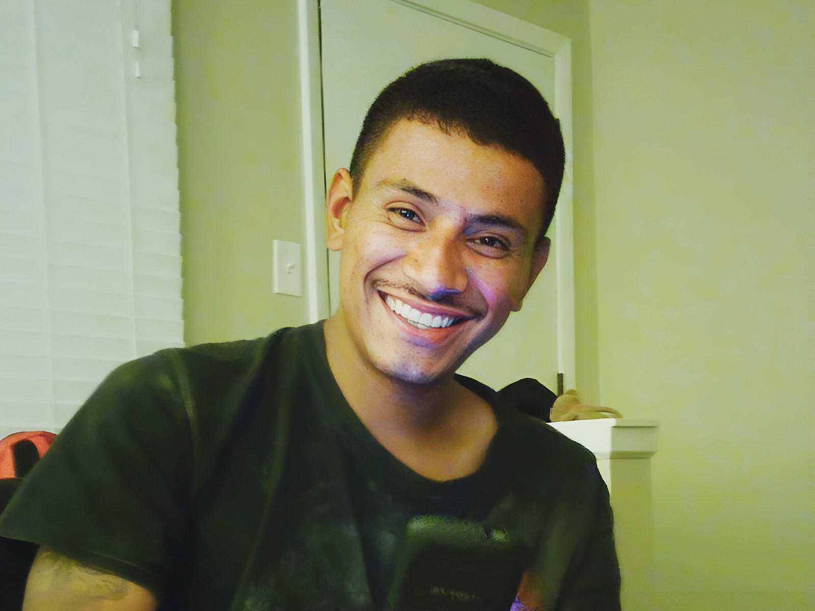 GreatKamil, smiling with short dark hair, wearing a black t-shirt, has a cute, attractive face with a small mustache and is standing in a well-lit room.