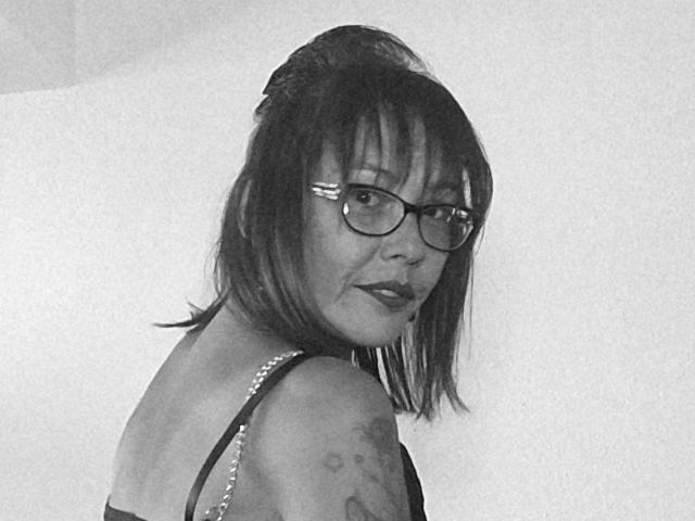 Black-and-white photo of LizzieStar69 with glasses, short dark hair, and a tattoo on her right shoulder. She's looking over her shoulder, wearing a strapless top.