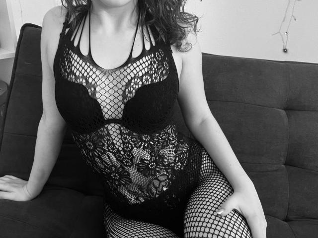 Sevillia in black fishnet lingerie, showing her boobs and pussy through the lace, sitting on a dark couch, arms resting on the seat. Black-and-white photo.