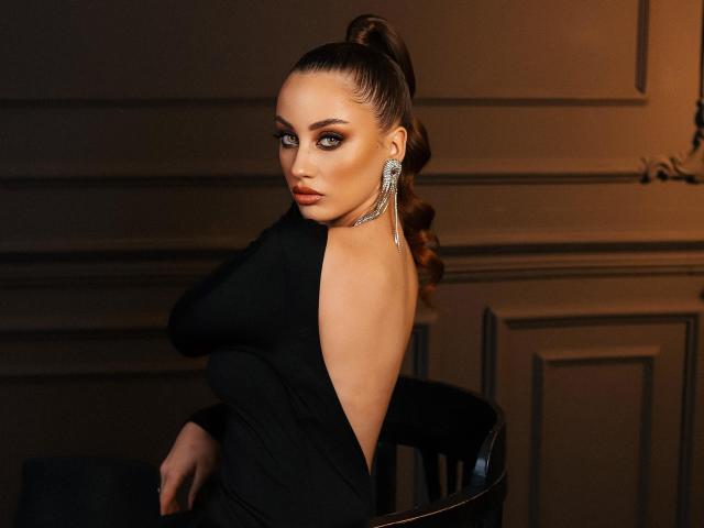 MaribelleVinnie with a high bun, wearing a black dress, showing her back with a low cut, looking over her shoulder, wearing dangly earrings, in a dimly lit room.