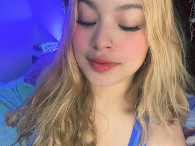 ShantalSummer with blonde hair, pink cheeks, and glossy pink lips, has long eyelashes, a nose ring, and a blue top, looking down with a slight smile.