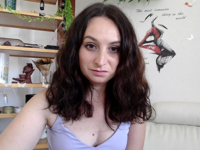 AskMeStandUp with wavy brown hair, light skin, and small tits in a purple tank top, looking directly at the camera with a slight smile. Shelves and wall art in background.