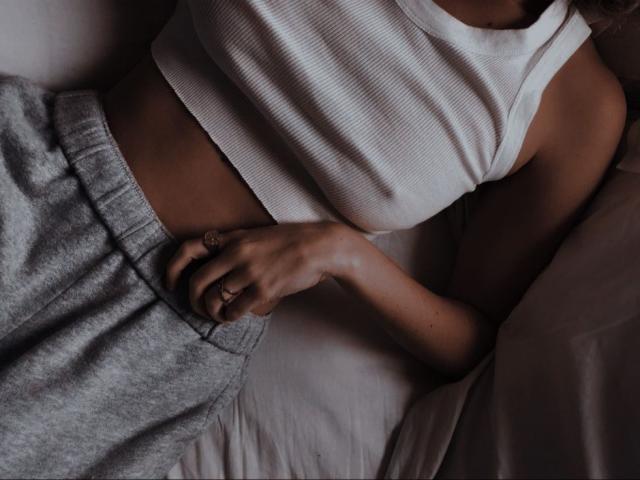 ChilliGold's tan skin, white ribbed tank top, and gray sweatpants. Her hand with a ring rests on her flat, toned belly. Her arm and chest are visible.