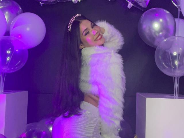 CarolineWill with long dark hair, smiling, wearing a fluffy white jacket, white striped pants, and a "Queen" crown, surrounded by purple-lit balloons. Her ass is prominent.