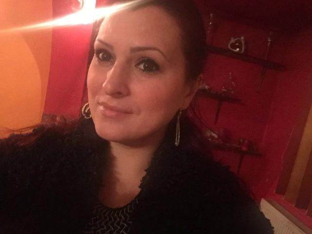SweetSonia-hot with brown hair, wearing a black fluffy coat, and silver earrings, has a slight smile and a light lens flare in a warmly lit red room.