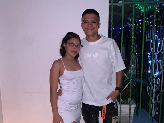 CouplePein with small tits in a white dress and CouplePein with a small butt in a white tee, standing together, smiling. He's wearing black pants, she's wearing glasses.