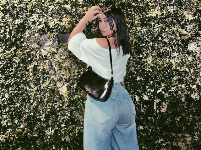 Lananexdoor15 with dark hair, white off-shoulder top, and blue high-waisted jeans, black shiny purse, holding sunglasses, looking back, showing her ass.