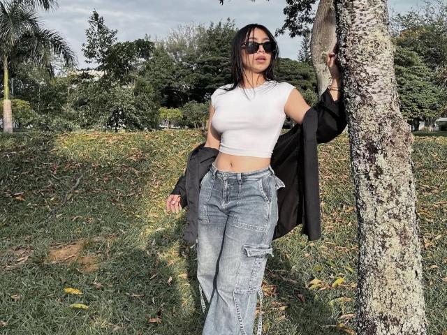 Lananexdoor15 stands by a tree, wearing a white crop top, blue jeans, and sunglasses, with a black jacket hanging off her arm, showing her small tits and slim ass.