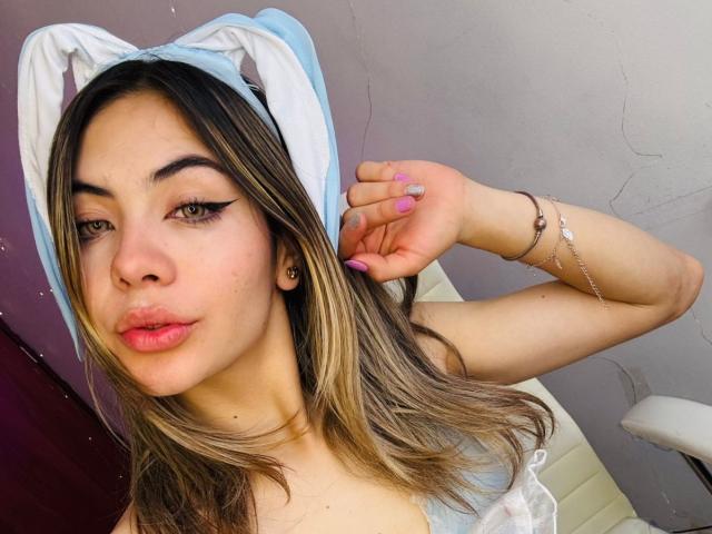 NathalySky with blonde hair, blue bunny headband, pink nails, winged eyeliner, pouty lips, light skin, small tits, arm raised, bracelet, slight crack in wall, white chair.