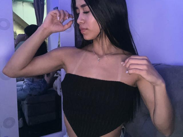 LaurentRay with long black hair, wearing a black strapless top, showing tan lines and small tits, standing with arms raised, in a purple-lit room, looking at a mirror.