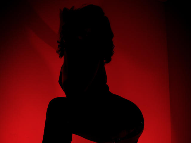 Silhouette of AdaMclain with curly hair, raised chin, and visible ass, against a red light, creating a dark, sexy, and mysterious effect. AdaMclain's tits are subtly outlined.