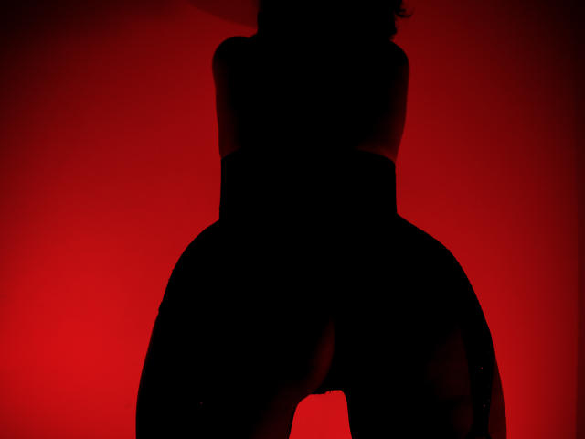 AdaMclain's black silhouette, butt and pussy prominently visible, against a bright red background, standing with her back to the camera, wearing garter straps, in a sexy, dark photo.
