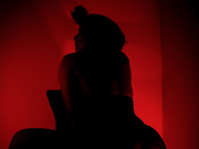 AdaMclain's back is to the camera, sitting with a raised butt, wearing a top, in a dark red, moody light. Her curly hair is in a bun.