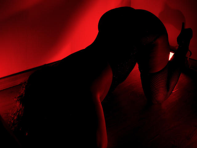AdaMclain is on all fours, wearing black fishnet stockings and a glittery dress, with red lighting highlighting her curvy ass and back, her curly hair partially visible.